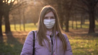 Beyaz tıbbi maskeli sevimli bir Avrupalı kızın portresi. Kız, akşam vakti parkta maske takarken kendini hasta hisseder. Solunum hastalıklarına karşı koruma. COVID-19. Salgına karşı küresel mücadele.