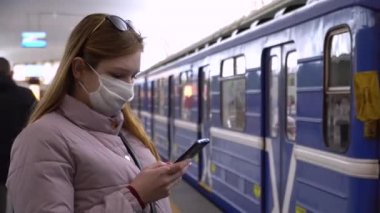 Maskeli bir kız metroda tıbbi maskeli bir akıllı telefon kullanıyor. Koronavirüs enfeksiyonunun önlenmesi. Enfeksiyonlara karşı solunum koruması. COVID-19