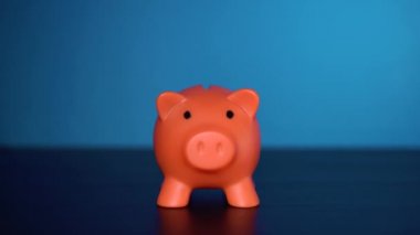Bir adamın eli kumbaraya bozuk para atıyor. Orange Piggy Bank mavi arka planda bir ışık masasının üzerinde duruyor. Yatırım yapmanın temelleri. Yatırım yapmak için güvenilir bir yol. Paranın değer kaybı..