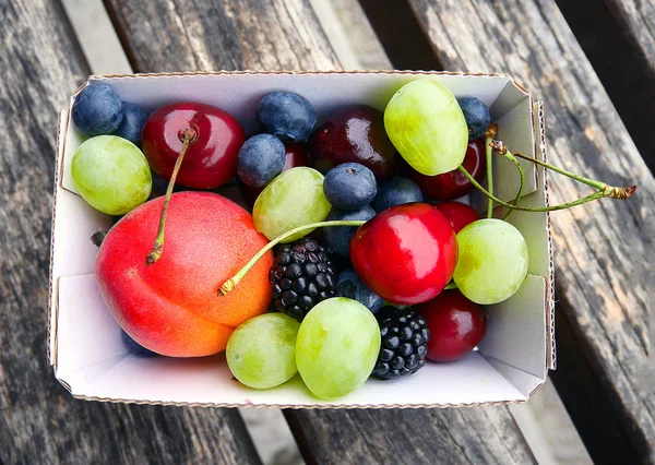 Mixed fruits in paper tray sold in Munich, Germany, including peach, green grapes, blueberries, blackberries, and cherry. The tray is set on a wood background