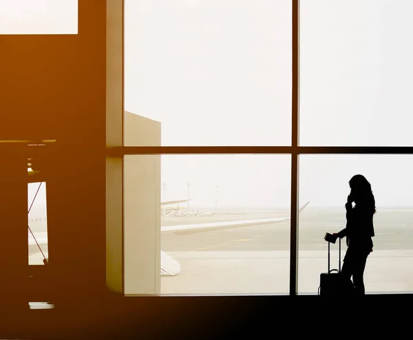 Silhouette del viaggiatore femminile che fa una telefonata di fronte al ponte jet nel cancello di partenza dell'aeroporto. Donna capelli lunghi ha portare con sé valigia, passaporto e carta d'imbarco. Dietro c'è una grande finestra e un aereo — Foto Stock