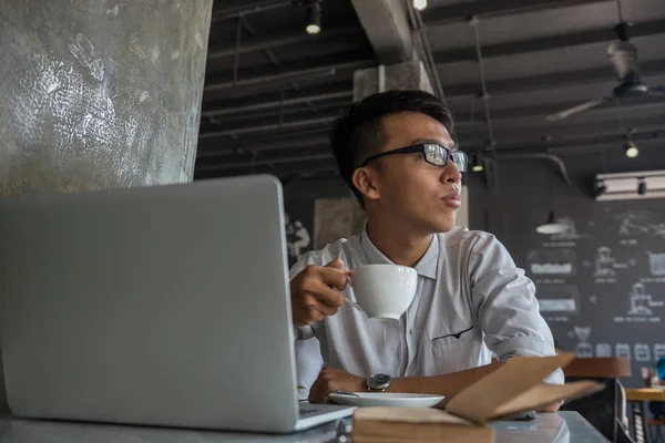 Joven hombre asiático sosteniendo taza de café y trabajando en el ordenador portátil — Foto de Stock