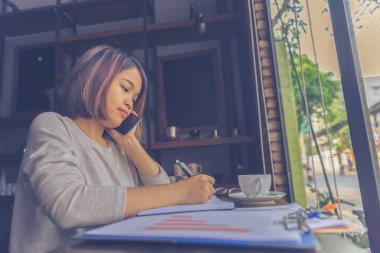 Genç Asyalı kadın defter yazarken cep telefonu kullanıyor.