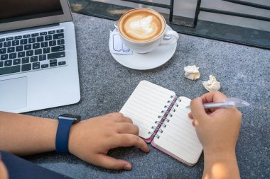 Laptopun ve sıcak kapuçinonun yanına notlar yazan bir kadın.