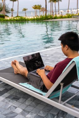Asyalı adam güzel yüzme havuzunda dizüstü bilgisayar kullanıyor.