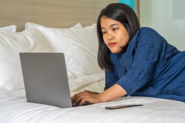 Genç bir kadın cep telefonunun yanında dizüstü bilgisayar kullanarak yatakta yatıyor.