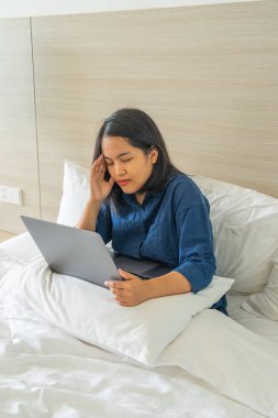 Baş ağrısı çeken Asyalı kadın yatak odasında dizüstü bilgisayarla çalışıyor.