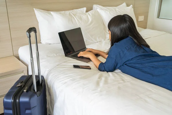 Femme asiatique couchée sur le lit tapant ordinateur portable à côté de la valise — Photo