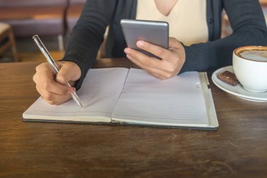 Genç Asyalı bayan akıllı telefon kullanıyor ve kafede notlar yazıyor.