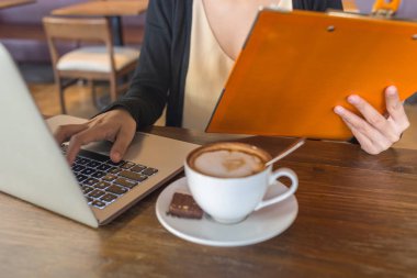 Dizüstü bilgisayarda yazan, elinde dosya tutan ve kahve içen genç bir kadın.