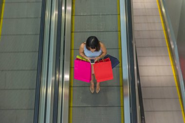 Yürüyen merdivende alışveriş torbaları tutan bir kadın akıllı telefon kullanıyor.