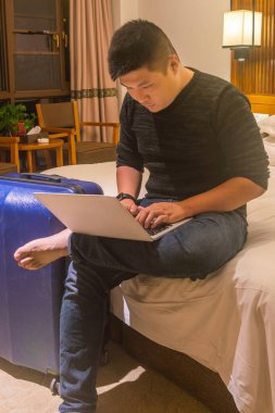 Adam yatakta oturuyor ve otel odasında dizüstü bilgisayar kullanıyor.