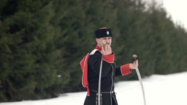 Молодой человек в казачьей одежде размахивает мечом в зимнем пейзаже на снегу . — стоковое видео