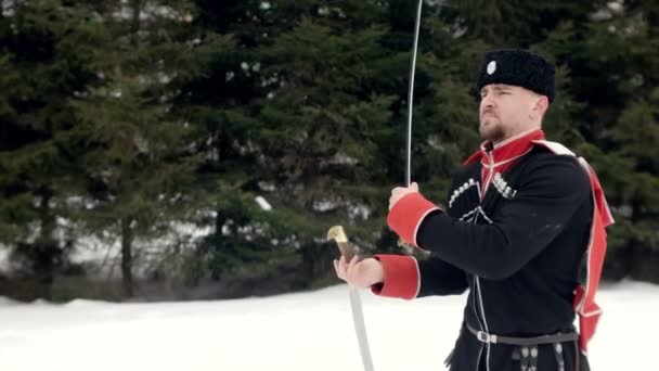 Un jeune homme en costume cosaque balançant une épée dans un paysage hivernal dans la neige . — Video
