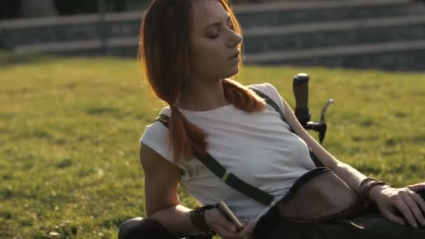 Рыжая женщина-велосипедистка отдыхает на зеленой лужайке в летнем парке — стоковое видео