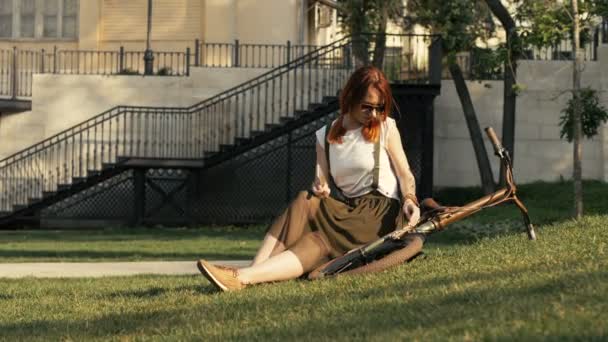 Unga rödhårig kvinna cyklist liggande på grönt gräs på city park i sommardag — Stockvideo