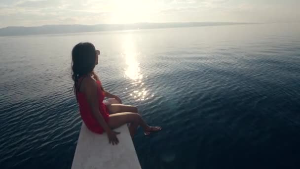 En flicka i röd klänning och solglasögon sitter på kanten av en båt. — Stockvideo