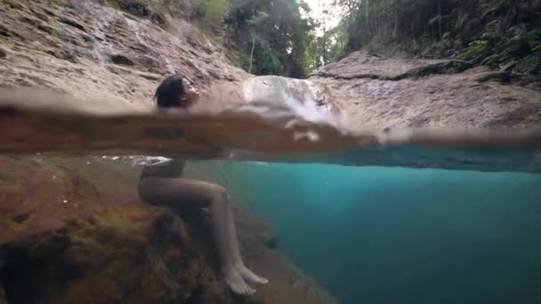 Mooie jonge vrouw zittend aan ongerepte waterval beek in het bos. — Stockvideo