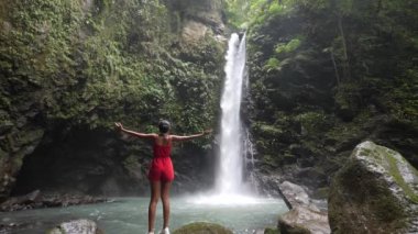 Slim young woman in a red romper raising hands in front of a scenic waterfall.