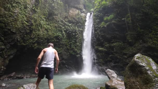 Male tourist standing at the foot of a waterfall amazed by the view of nature. — Stockvideo