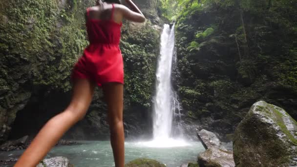 Молодая женщина в красном комбинезоне ходит и прыгает у подножия водопада . — стоковое видео