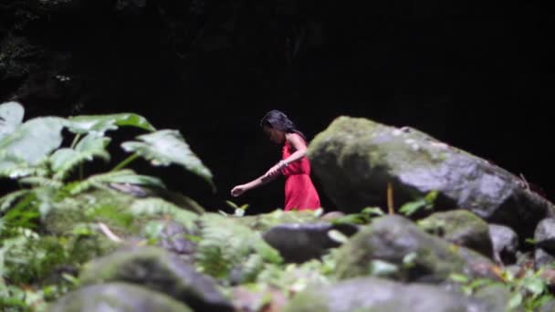 Νεαρή γυναίκα με κόκκινο φόρεμα περπατά μόνη κοντά σε μια σκοτεινή σπηλιά στη ζούγκλα. — Αρχείο Βίντεο