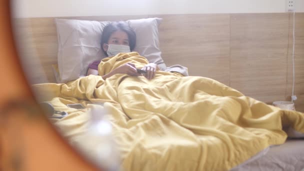 En sjuk ung kvinna som ligger i sängen och bär mask och tittar på nyheter på TV. — Stockvideo