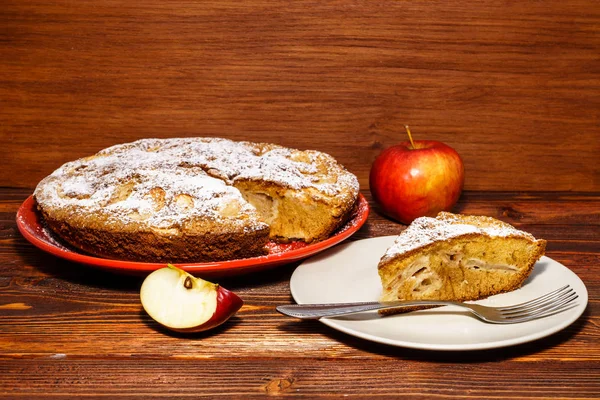 Tarta de manzana recién horneada sobre un fondo rústico de madera oscura. Manzanas frescas y un trozo de tarta de manzana espolvoreada con azúcar en polvo en el fondo de una tabla oscura . — Foto de Stock