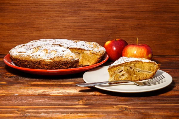 Tarta de manzana recién horneada sobre un fondo rústico de madera oscura. Manzanas frescas y un trozo de tarta de manzana espolvoreada con azúcar en polvo en el fondo de una tabla oscura . — Foto de Stock