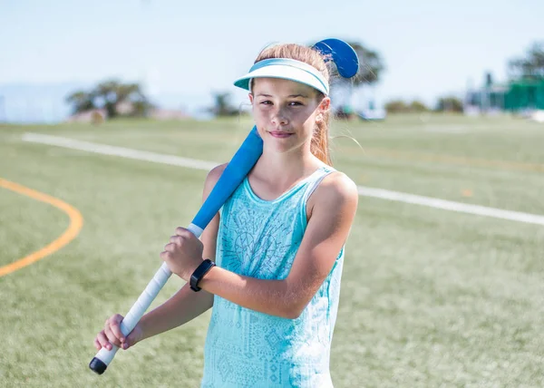 Sporty girl standing on sports field holding a blue hockey stick looking at the camera smiling.