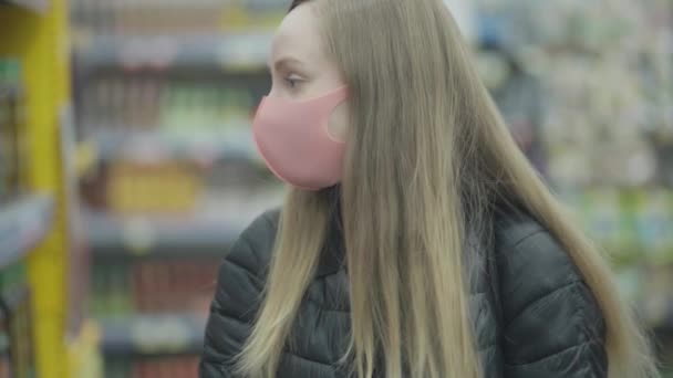 Covid-19-sepelvaltimovirus, pandemia. vakava nuori blondi tyttö seisoo naamio käsineet ostoskorin supermarketissa taustaa vasten asiakkaiden, kameran liikkeen — kuvapankkivideo