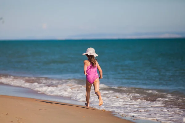 Muchacha joven en traje de baño y sombrero corre a lo largo de la línea de surf en una playa de arena —  Fotos de Stock