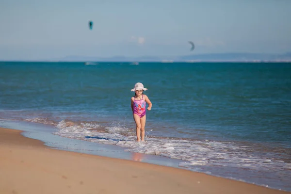 Muchacha joven en traje de baño y sombrero corre a lo largo de la línea de surf en una playa de arena —  Fotos de Stock