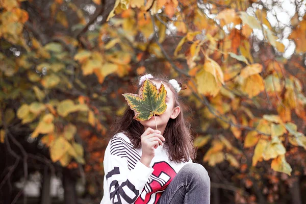 Beautiful little girl holds an autumn leaf in her hand and enjoys the autumn. Autumn, backgrounds, close-up.Girl holding colorful leaf in autumn park. — Stock Photo, Image