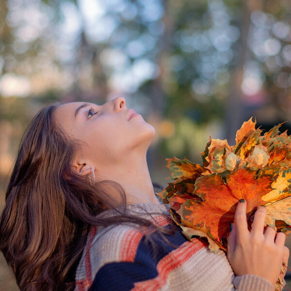 Beautiful girl sits on a bench in autumn. In the girl's hand holds autumn leaves. girl sitting at bench in autumn park.