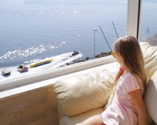 Een klein meisje zit bij het raam en kijkt naar de oceaan. Het uitzicht vanuit het raam naar de zee.Achteraanzicht van jonge vrouw met uitzicht op zee vanaf balkon in resort. — Stockfoto