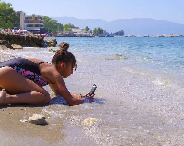 Młoda piękna dziewczyna siedzi na plaży w pobliżu morza i robi zdjęcia na telefon.Sezon letni. Morze i plaża. — Zdjęcie stockowe