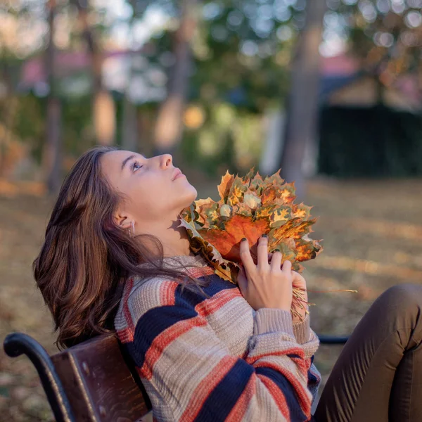 一个小女孩坐在街上的长椅上，手里拿着秋天的叶子。 秋天的概念。 美丽的秋天. — 图库照片