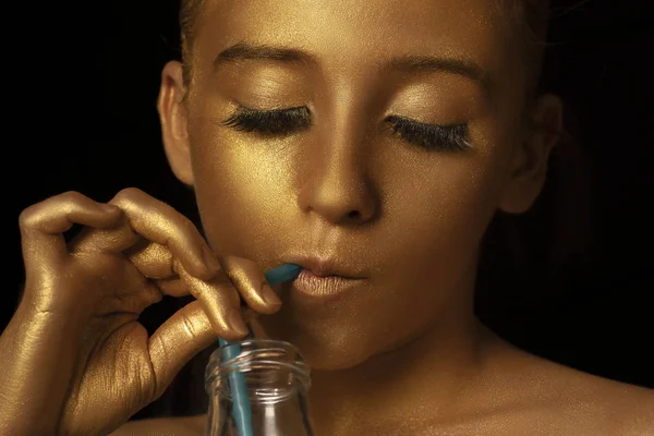 Gold Fashion Model Beauty Portrait with a bottle and a cocktail straw , Golden Woman Art Makeup on studio black background.Beauty woman face makeup close up.