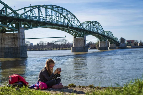 Una niña está sentada en la orilla del río con un teléfono. En un cálido día de primavera . — Foto de Stock