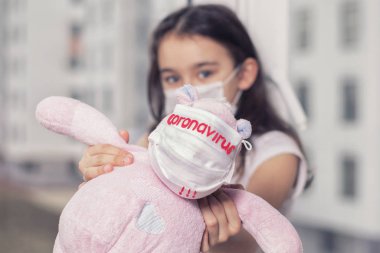 Yüzünde virüsten koruyucu bir maske olan küçük bir kız, koruyucu maskeli bir oyuncakla pencerenin kenarında oturur, Coronavirus yazısıyla, kız üzülür. Kişisel koruyucu ekipman gereklidir..