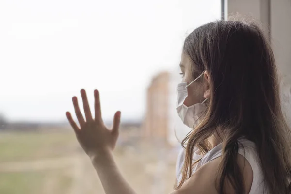 Çin 'deki koronavirüs salgını. Coronovirus Karantina Konsepti. Covid-19 'u korumak için maske takan küçük kız pencerenin yanında duruyor ve üzgün. Kız hastalandı ve dışarı çıkamıyor..