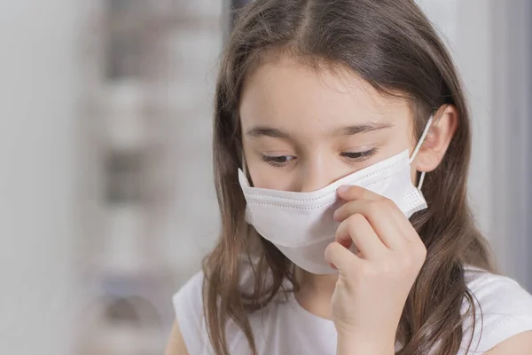 Çin 'deki koronavirüs salgını. Coronovirus Karantina Konsepti. Covid-19 'u korumak için maske takan küçük kız pencerenin yanında duruyor ve üzgün. Kız hastalandı ve dışarı çıkamıyor..