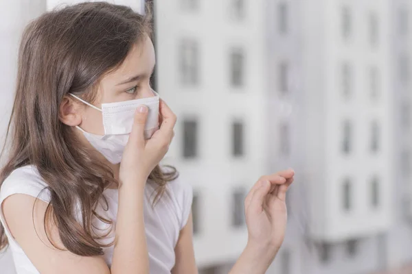 Çin 'deki koronavirüs salgını. Coronovirus Karantina Konsepti. Covid-19 'u korumak için maske takan küçük kız pencerenin yanında duruyor ve üzgün. Kız hastalandı ve dışarı çıkamıyor..