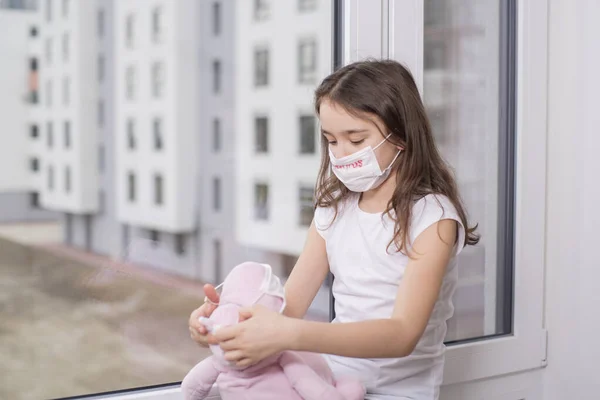 Yüzünde virüsün koruyucu maskesi olan beyaz tişörtlü küçük bir kız elinde oyuncakla pencerenin kenarında oturuyor ve üzülüyor. Korumalı maskeli, Coronavirus yazılı bir oyuncak taşıyan bir kız..