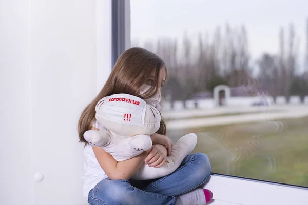 Yüzünde virüsün koruyucu maskesi olan beyaz tişörtlü küçük bir kız elinde oyuncakla pencerenin kenarında oturuyor ve üzülüyor. Korumalı maskeli, Coronavirus yazılı bir oyuncak taşıyan bir kız..