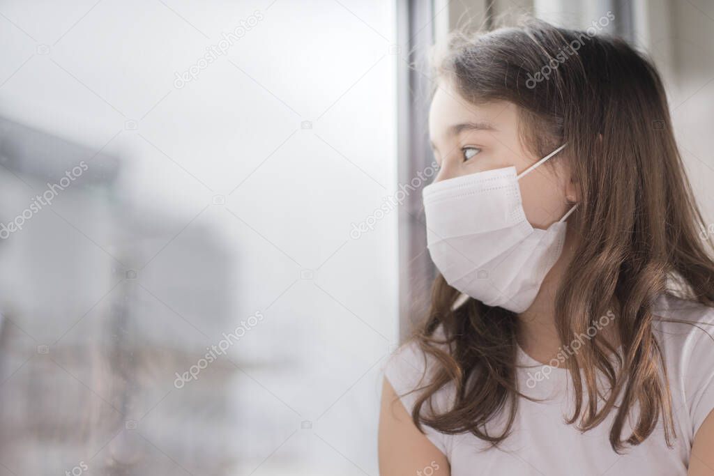 The Chinese outbreak of coronavirus. Coronovirus Quarantine Concept.Little girl wearing mask for protect Covid-19 stands near the window and is sad. The girl became ill and cannot go out.