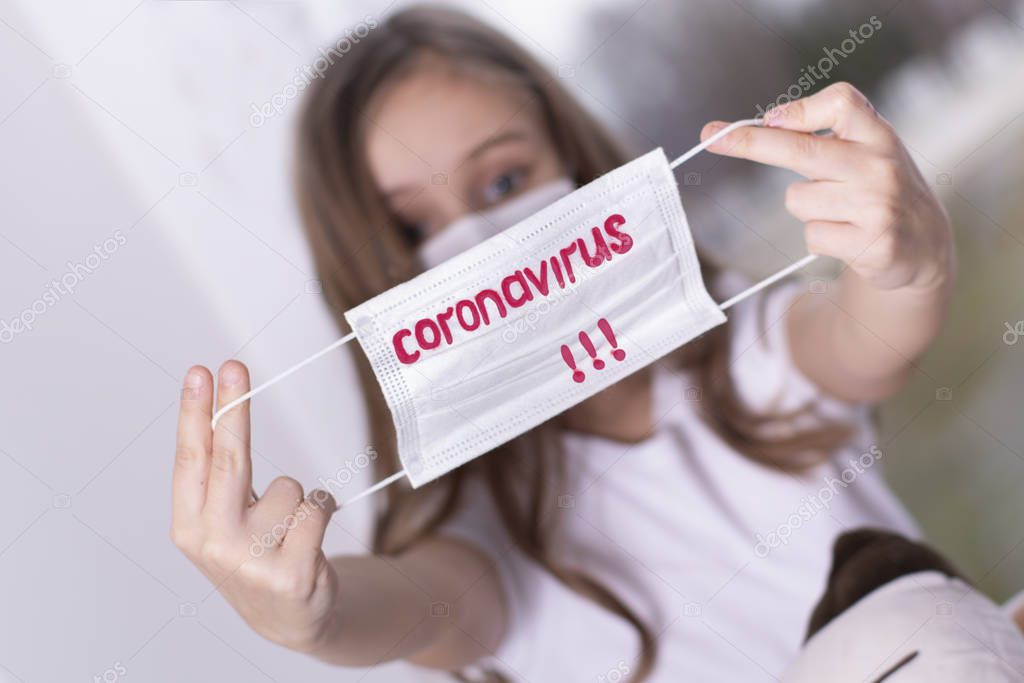 Coronovirus Quarantine Concept.Personal Little girl is sitting by the window in her hands a protective mask with the text Coronavirus, girl is sad. Girl in a protective mask by the window.
