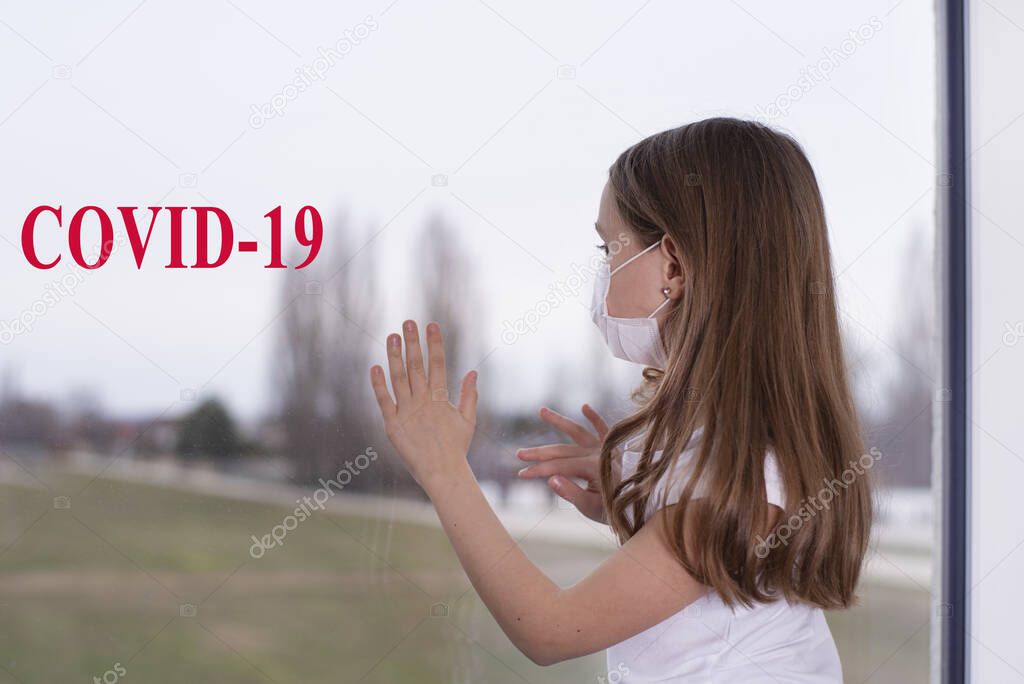 A little girl sits at the window in a protective mask, wants to go outside, but it s impossible, there is a coronavirus, the girl is sad.Coronovirus Quarantine Concept.