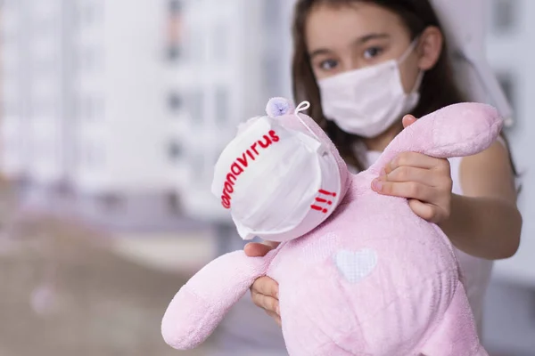 Yüzünde virüsten koruyucu bir maske olan küçük bir kız, elinde koruyucu bir maskeyle pencerenin kenarında oturuyor, Coronavirus yazısıyla, kız üzgün. Wuhan coronavirüs ve salgın virüsü belirtileri gösteriyor..